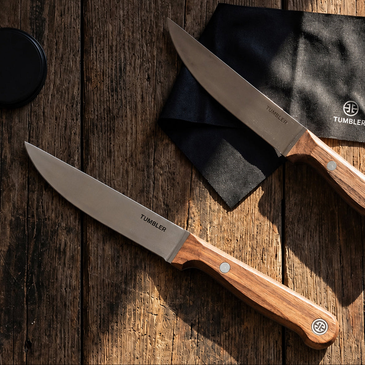 Two knives with wooden handles on a wooden surface, featuring the Tumbler brand.
